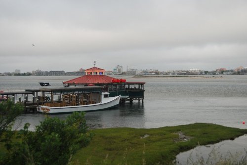 A boat docked at a waterfront restaurant under a cloudy sky, with a city skyline in the background.