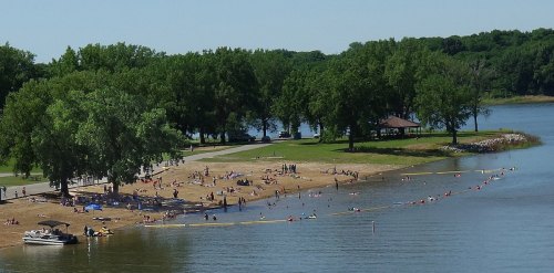 A sunny lakeside scene with people swimming and relaxing on the beach, surrounded by trees and a calm water view.