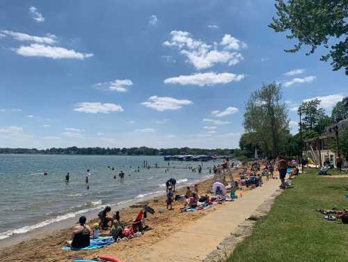 A sunny beach scene with people relaxing on the sand, swimming, and enjoying the water under a clear blue sky.