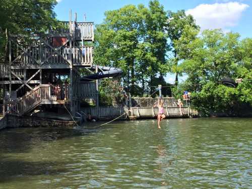 A wooden structure by a lake with people enjoying activities, including a girl on a rope swing over the water.