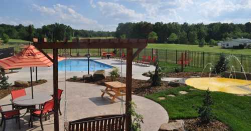 A sunny outdoor pool area with lounge chairs, a picnic table, and a splash pad, surrounded by greenery and trees.