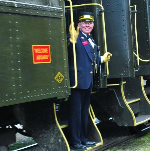 A smiling train conductor waves from the doorway of a vintage train car, with a "Welcome Aboard" sign visible.