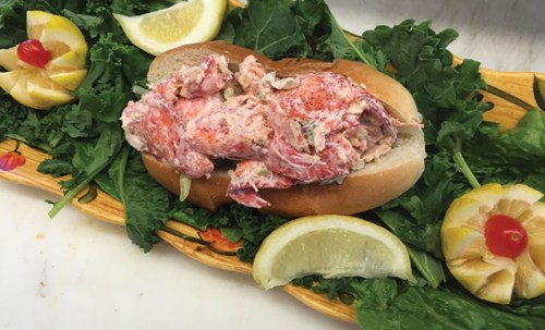 A lobster roll filled with lobster meat, served on a bed of greens, garnished with lemon wedges and cherry tomatoes.
