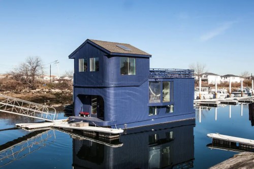 A blue houseboat with a two-story design docked at a marina, reflecting in calm water under a clear sky.