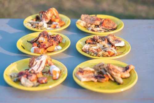 Six yellow plates with grilled chicken, vegetables, and sauces arranged on a table outdoors.