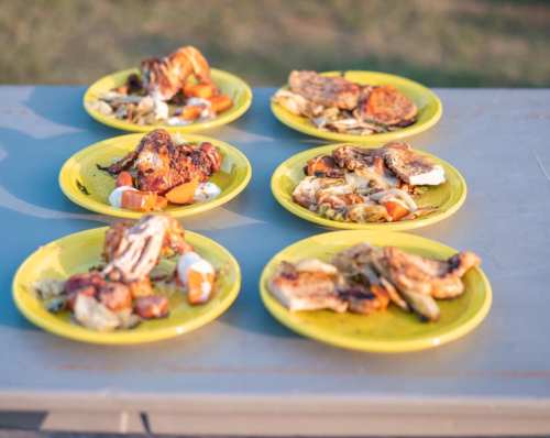 Six yellow plates with grilled chicken and assorted vegetables, arranged on a table outdoors.