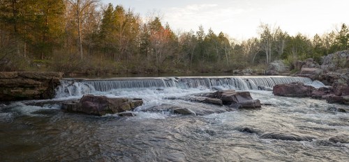 A serene river scene with a waterfall cascading over rocks, surrounded by lush trees and a clear sky.