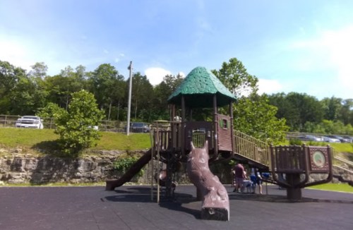A playground structure with a green roof, slides, and climbing features, surrounded by trees and parked cars in the background.