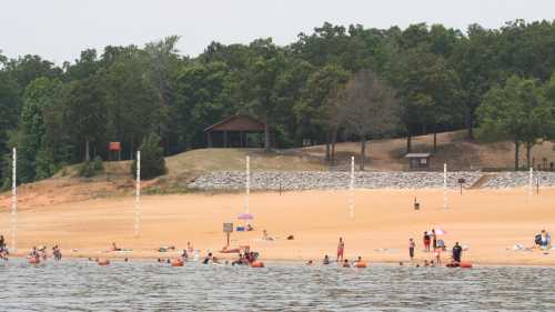 A sandy beach with people swimming and relaxing, surrounded by trees and a pavilion in the background.