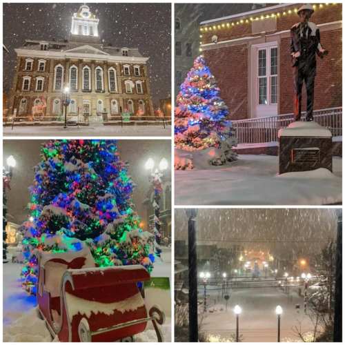 A snowy town square featuring a historic building, decorated Christmas trees, a statue, and a festive sleigh.