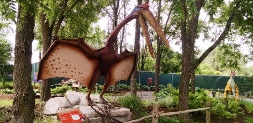 A large, colorful dinosaur sculpture with wings stands among trees in a park, surrounded by greenery.