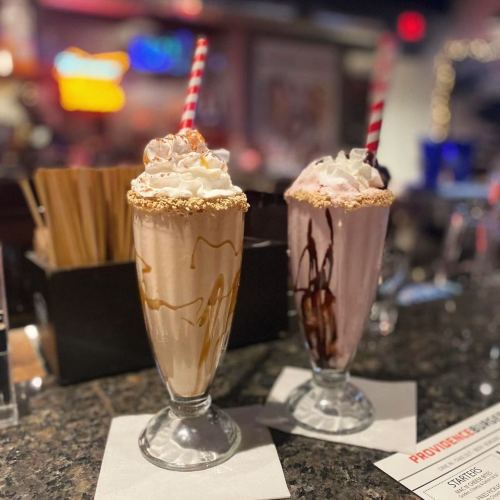 Two tall milkshakes in glasses, one topped with whipped cream and caramel, the other with chocolate and a cherry, on a bar counter.