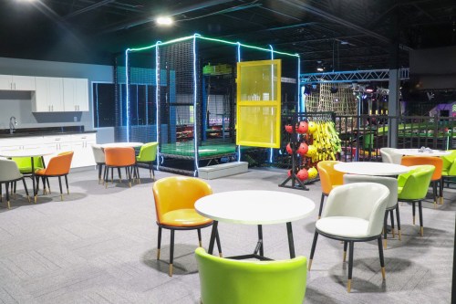 A colorful indoor play area featuring tables and chairs, with a netted play structure in the background.