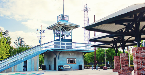 A modern train station with a raised platform, surrounded by greenery and a clear blue sky.