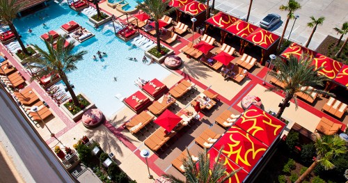 Aerial view of a vibrant pool area with lounge chairs, cabanas, and palm trees, filled with people enjoying the sun.