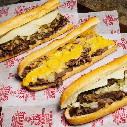 Three delicious cheesesteak sandwiches on paper, featuring beef, cheese, and toppings like onions in fresh hoagie rolls.