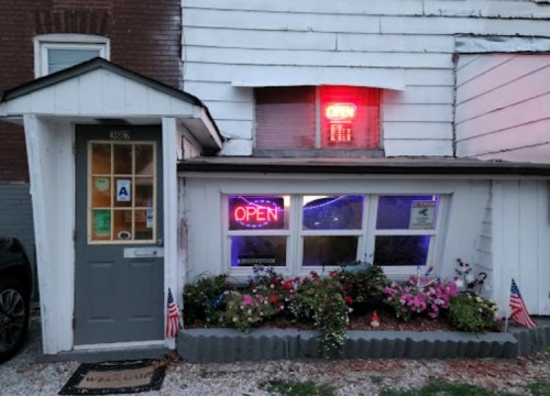 A small building with an "OPEN" neon sign, colorful flowers, and American flags in front.