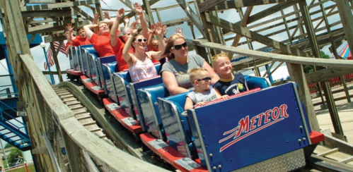 A group of excited people on a roller coaster, smiling and raising their hands, with a wooden structure in the background.