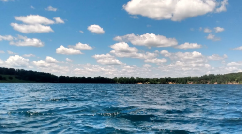 A serene lake under a blue sky filled with fluffy white clouds, surrounded by green trees on the horizon.