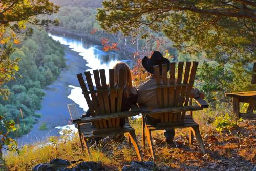 A couple sits on wooden chairs overlooking a river at sunset, surrounded by trees and nature.
