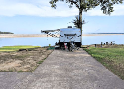 A camper parked by a calm lake, with bicycles nearby and a picnic area in the background under a clear sky.