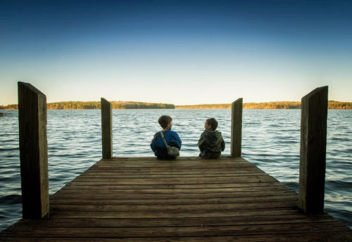 Two children sit on a wooden dock, gazing out at a calm lake surrounded by trees under a clear blue sky.