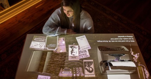 A woman sits at a table with a typewriter, surrounded by books and papers, with projected text illuminating the scene.