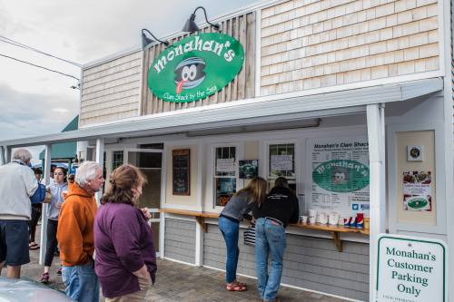 A busy clam shack with a sign, people ordering at the counter, and a few customers waiting outside.