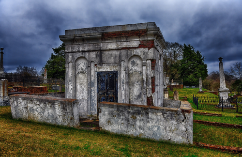A weathered mausoleum in a cemetery, surrounded by gravestones and overcast skies.