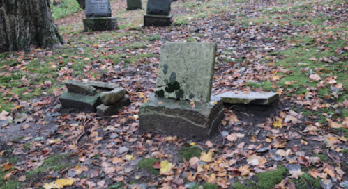 A weathered gravestone surrounded by fallen leaves and scattered stones in a quiet, overgrown cemetery.