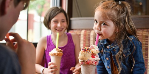 Two girls enjoy colorful milkshakes with whipped cream and sprinkles, smiling and having fun in a cozy setting.