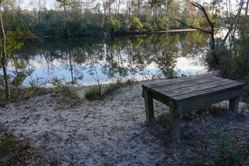 A wooden bench by a calm river, surrounded by trees and sandy ground, reflecting the serene landscape.
