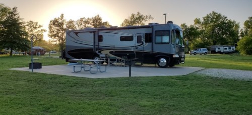 A parked RV on a concrete pad in a grassy area, with trees and a sunset in the background.