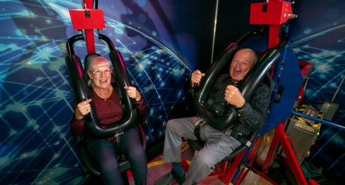 Two elderly individuals sit in amusement ride seats, smiling and holding onto safety harnesses, ready for a thrill.