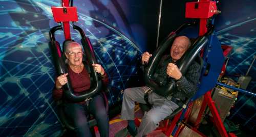 Two elderly individuals sit in amusement ride seats, smiling and holding onto safety harnesses, ready for a thrill.