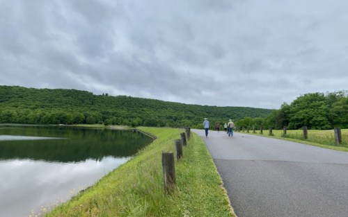 Two people walk along a path beside a calm lake, surrounded by lush green hills under a cloudy sky.