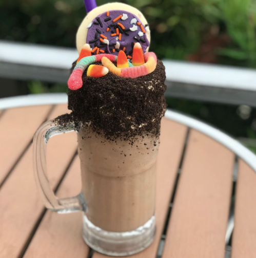 A tall milkshake topped with cookie, sprinkles, gummy worms, and chocolate crumbs, served in a clear glass.