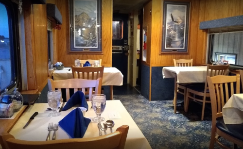 Cozy restaurant interior with wooden decor, tables set with blue napkins, and large windows showing a scenic view.