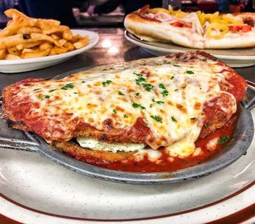 A plate of chicken parmesan topped with melted cheese and marinara sauce, served with fries in the background.