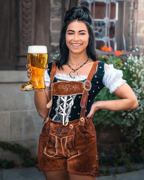 A woman in traditional Bavarian attire holds a large beer mug, smiling in front of a rustic building with flowers.