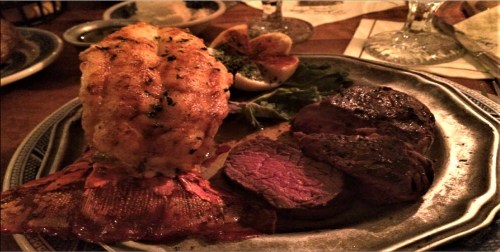 A plate featuring a lobster tail and slices of tender beef, garnished with herbs and lemon wedges.