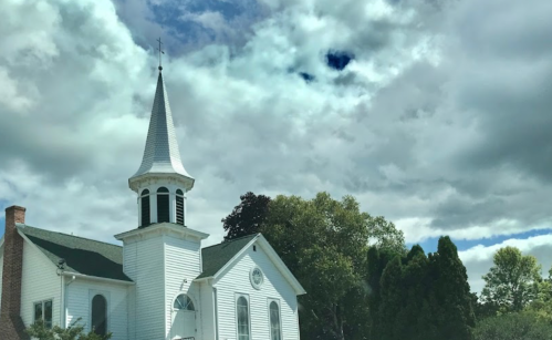 A white church with a tall steeple surrounded by trees under a cloudy sky.