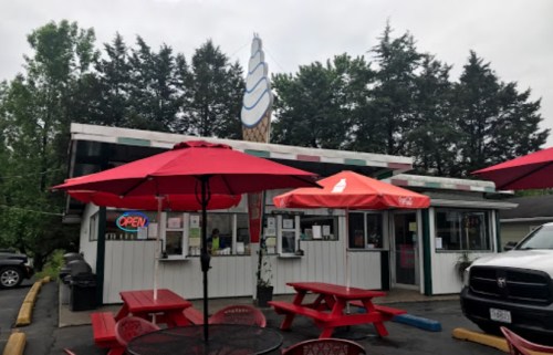 A small ice cream shop with red umbrellas, picnic tables, and a large ice cream cone sign, surrounded by trees.