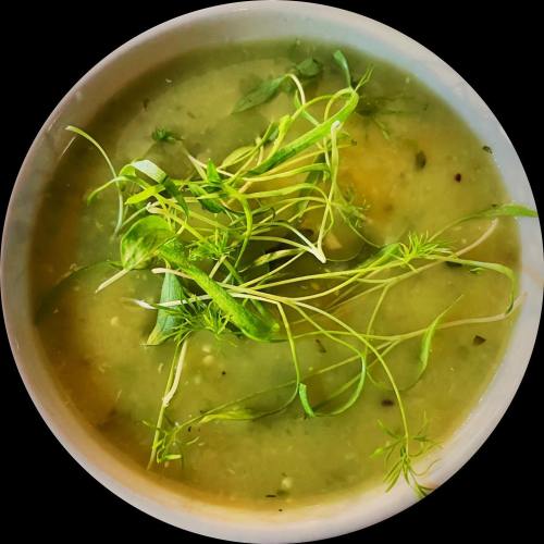 A bowl of green soup garnished with fresh herbs and pea shoots, set against a black background.