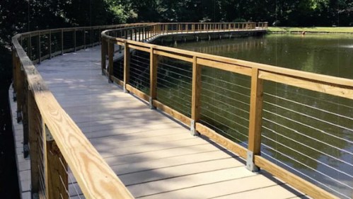 A curved wooden boardwalk with a railing, extending over a calm pond surrounded by trees.