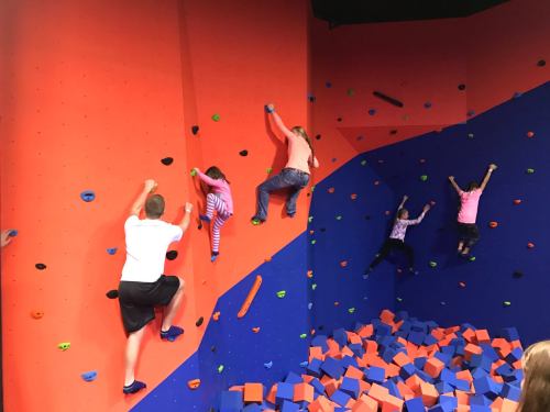 A group of children and an adult climbing a colorful indoor rock wall with foam blocks below.