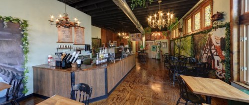 Cozy café interior with wooden tables, a counter, and decorative murals of nature and buildings. Chandeliers hang from the ceiling.