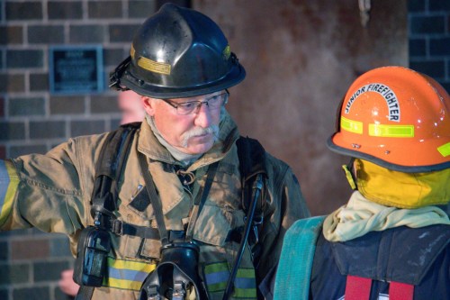 A firefighter in gear talks to a junior firefighter, both wearing helmets, in a training environment.