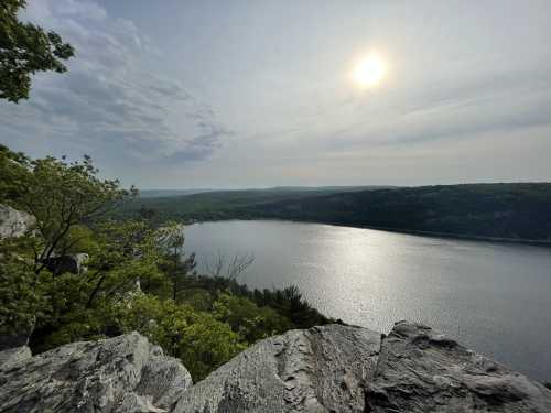 A serene view of a river surrounded by lush greenery and rocky cliffs under a cloudy sky with the sun shining.