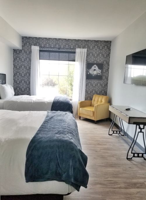 A modern hotel room with two beds, a yellow armchair, and a large window with natural light.
